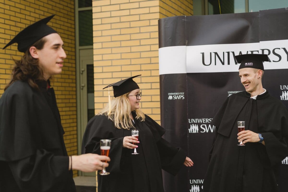 Three graduates wearing black caps and aprons are celebrating outside near a black and white "University Swaps" banner. Holding glasses of what appears to be sparkling wine, they are engaged in animated conversation, and one of the graduates is gesticulating animatedly. It's a perfect photo recap of the events capturing their joy.  