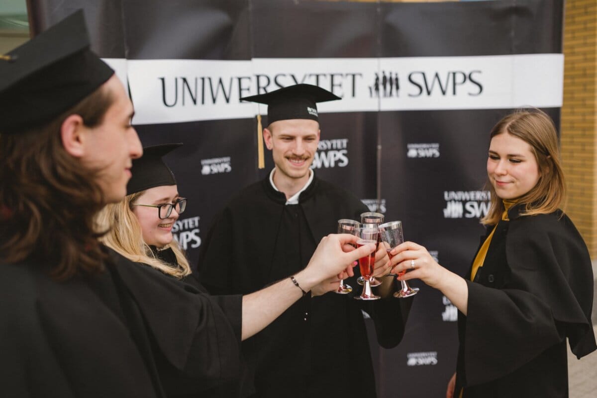 Four graduates of academic outfits celebrate by clinking their glasses against the backdrop of the "SWPS UNIVERSITY" sign. They are smiling and seem happy, completing their academic journey, which was perfectly captured by Marcin Krokowski, a well-known event photographer Warsaw. 