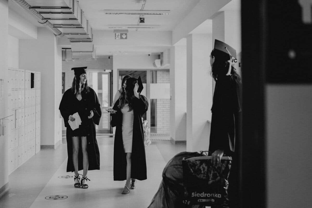 Black and white photo of three people walking down a corridor wearing graduation caps and aprons. Two are walking together, one is holding a piece of paper, the third is standing a few steps away with a suitcase. Captured by an event photographer in Warsaw, it captures the moment perfectly.  