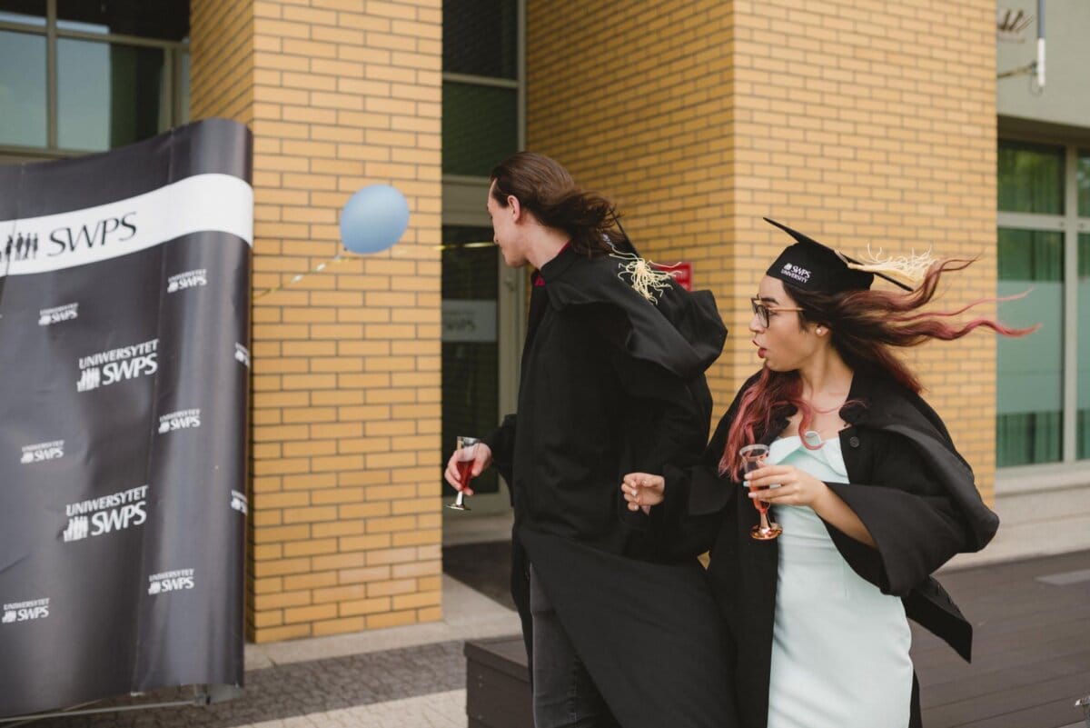 Two graduates in black aprons and caps hold drinks and walk past the scene with "SWPS University." The wind blows away the hair of the woman on the right, and a blue balloon floats past her. They seem surprised, perhaps reacting to something in this event photo.  