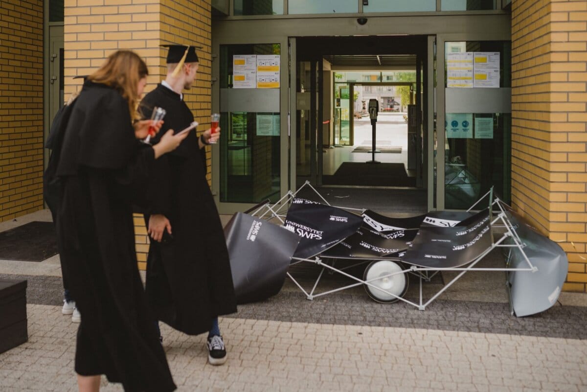 Two people in aprons and graduation caps stand in front of the building. They look at the collapsed banner frame on the ground near the entrance and seem amused. The building has yellow brick walls and glass doors with signage - a perfect example of Marcin Krokowski's event photography.  