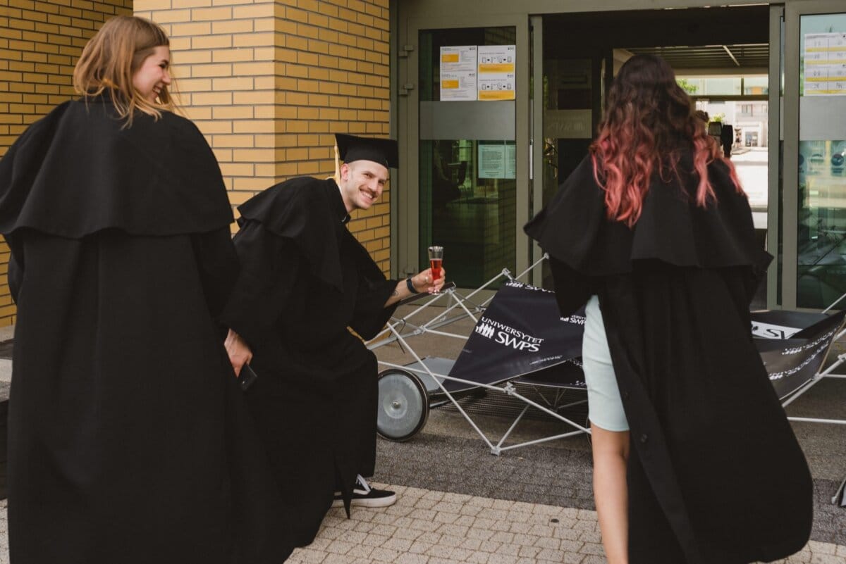 Three people in graduation togas stand in front of the building. One person crouches, smiles and holds a drink next to a broken umbrella cart. Two others are walking toward the building. Next to them on the cart is a stack of posters - a perfect scene for an event photographer Warsaw.   