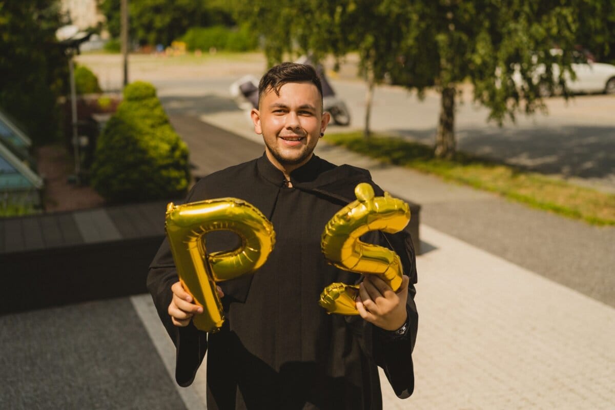 A person wearing a graduation gown and holding gold foil balloons in the shape of the letters "P" and "S." They are outside on a sunny day, with greenery and a parking lot in the background. The person is smiling, capturing the joy of this special day in a photo essay from Marcin Krokowski's event.  