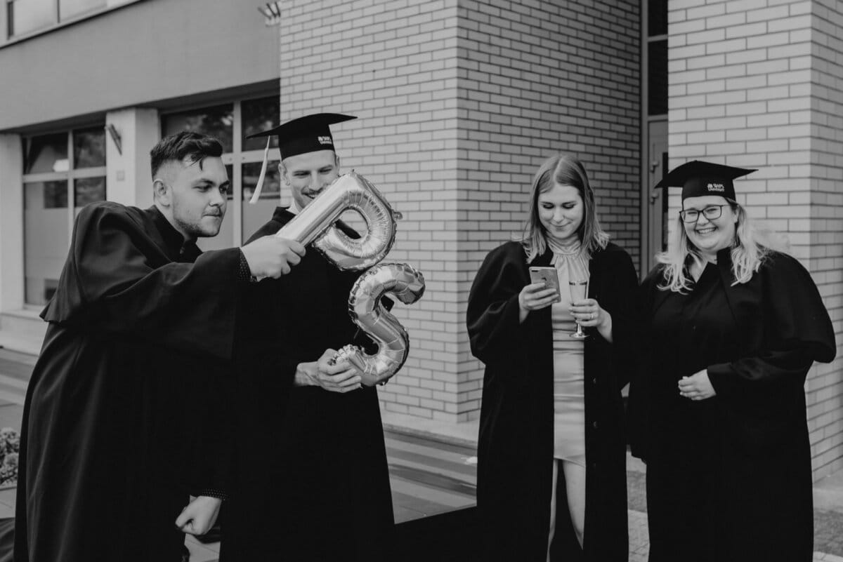 The black-and-white photo shows four people in aprons and graduation caps standing near a brick building. One is holding a balloon with the letter "S," while another appears to be capturing the moment on his phone. The group gives the impression of being joyful and celebratory, perfect for a photo essay of the event.  