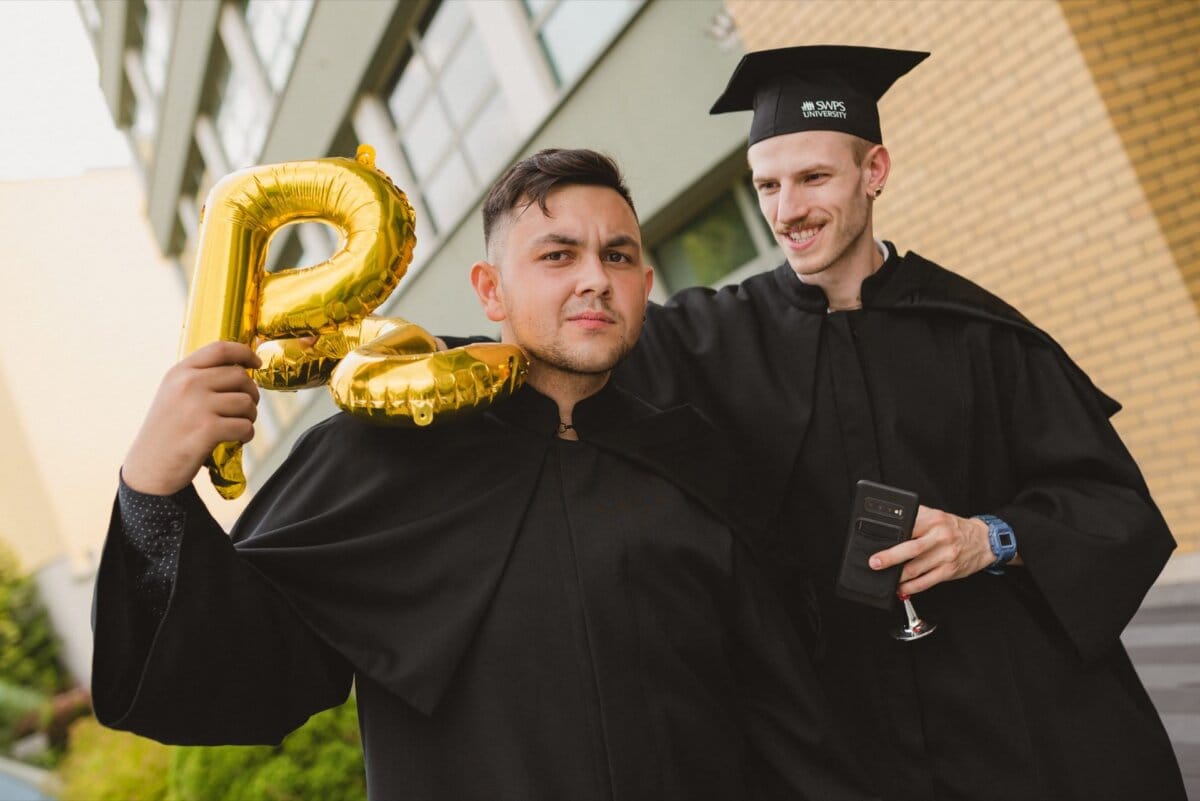 Two men in caps and togas celebrate. The man on the left holds a gold balloon shaped like a "B" with a neutral expression on his face, while the man on the right smiles, holding a smartphone in one hand and embracing a friend. Capture this joyful moment with an event photographer to preserve lasting memories.  