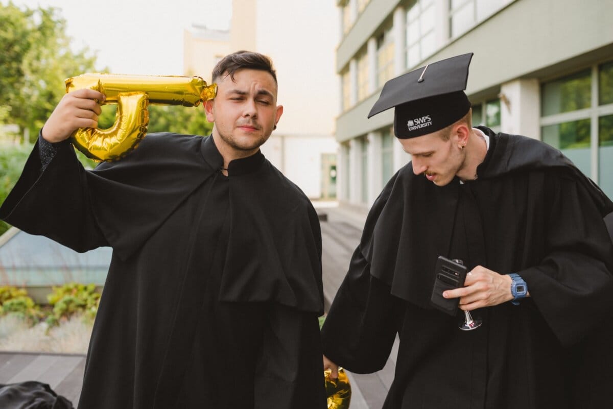 Two young men in aprons and end-of-year caps are celebrating outdoors. One holds an inflated gold balloon in the shape of a number near his head, while the other looks at his phone and holds a small wine glass. In the background is a modern building and greenery, perfectly captured by Marcin Krokowski.  