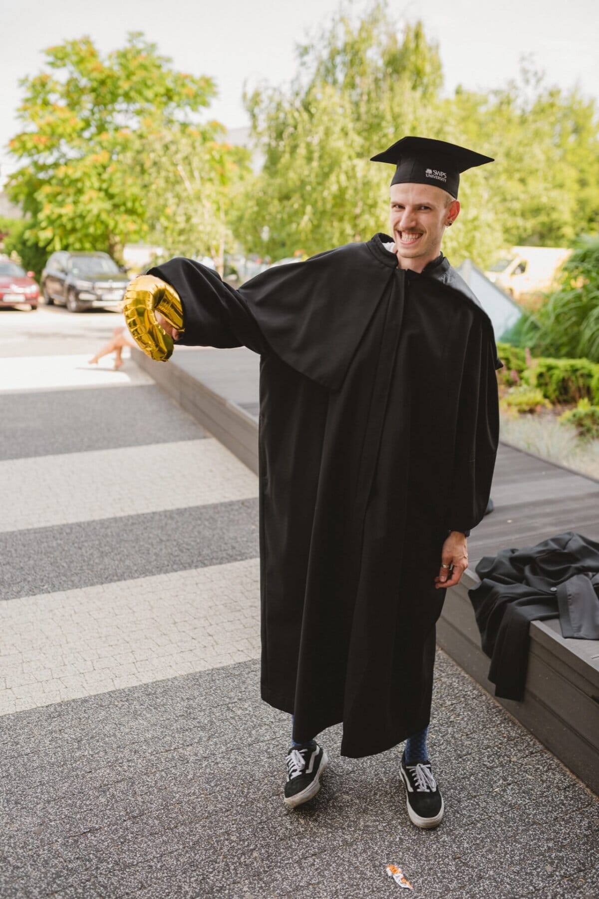 A person wearing a graduation cap and gown is standing on a paved area outside. They are holding a golden balloon in the shape of the number one and smiling excitedly. In the background you can see trees and parked cars. This photo shows a great event photo by a talented event photographer.   