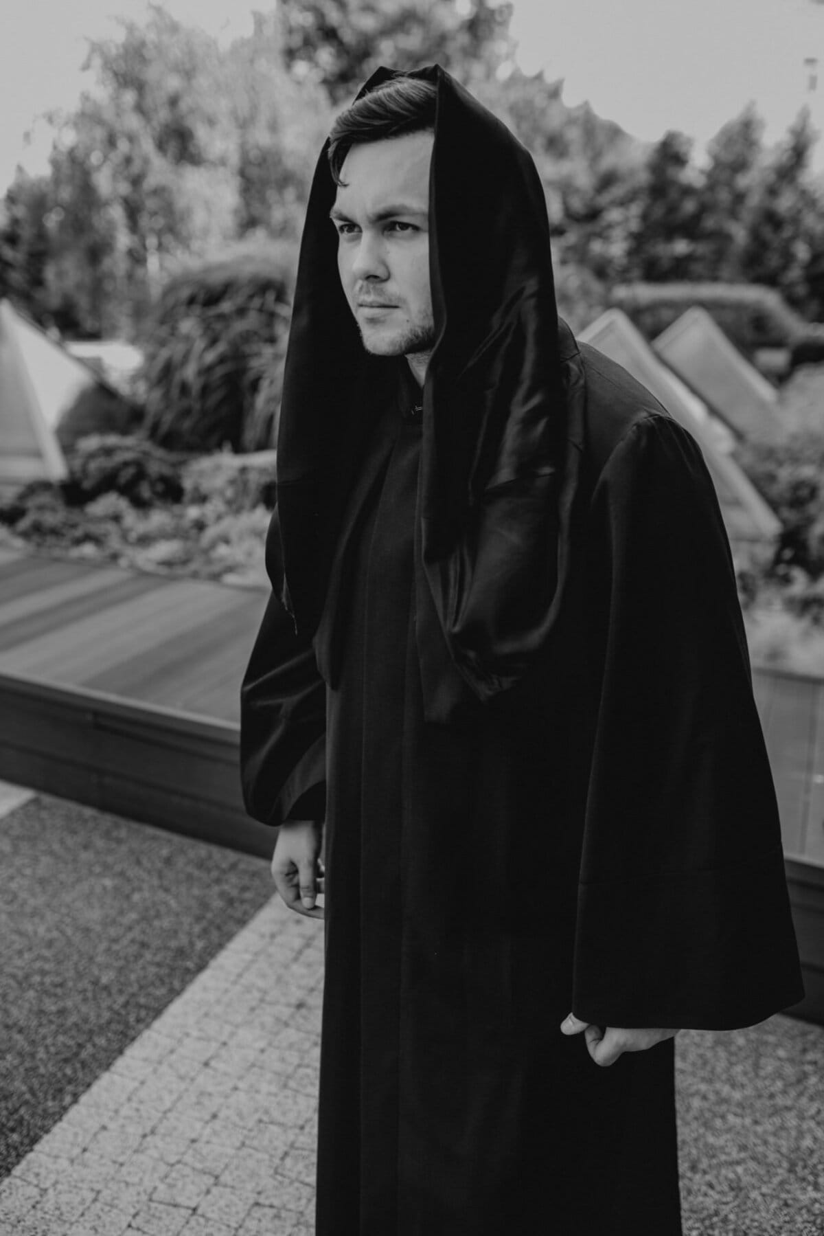 A person dressed in a dark robe with a hood stands outside in a garden-like area. The black and white photo, taken by Marcin Krokowski, emphasizes the mysterious and serious expression on their faces. Geometric structures and leaves are visible in the background, adding to the enigmatic atmosphere.  