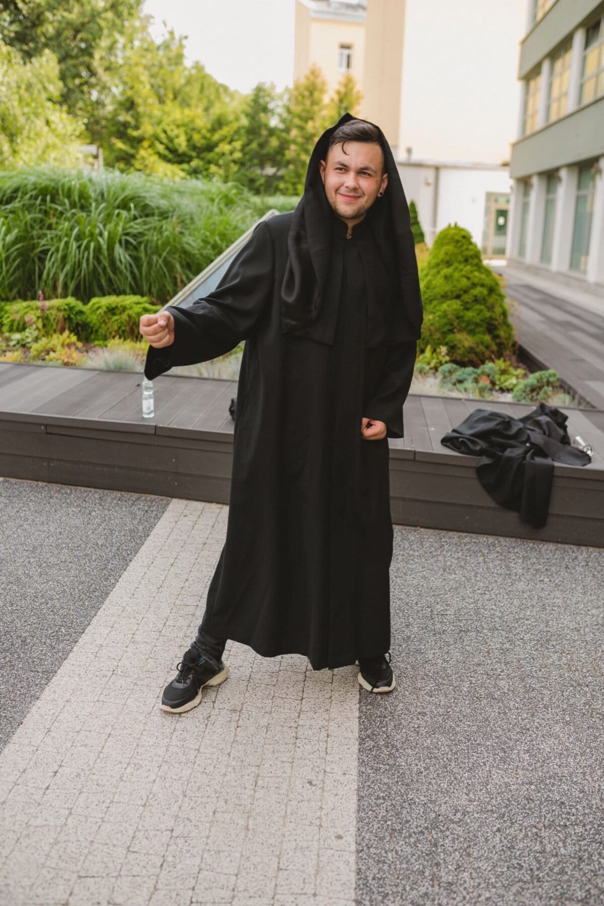 The person is outside wearing a long black robe with a hood. They are standing on a cobblestone path surrounded by greenery and a building in the background. The person, possibly part of Marcin Krokowski's event photography, makes a playful face and points forward with his right hand.  