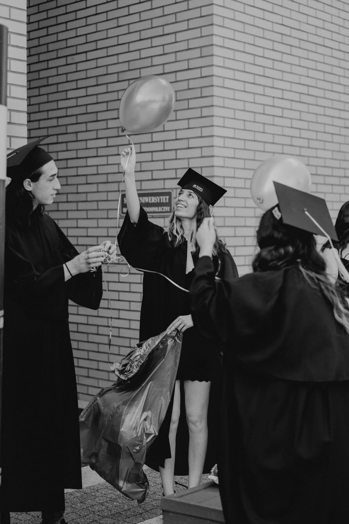 A group of graduates in caps and aprons stand together against a brick wall. One graduate, smiling radiantly, holds a balloon while another prepares to inflate it. They are celebrating, and the mood is joyous and festive, capturing a wonderful photo report of the events.  