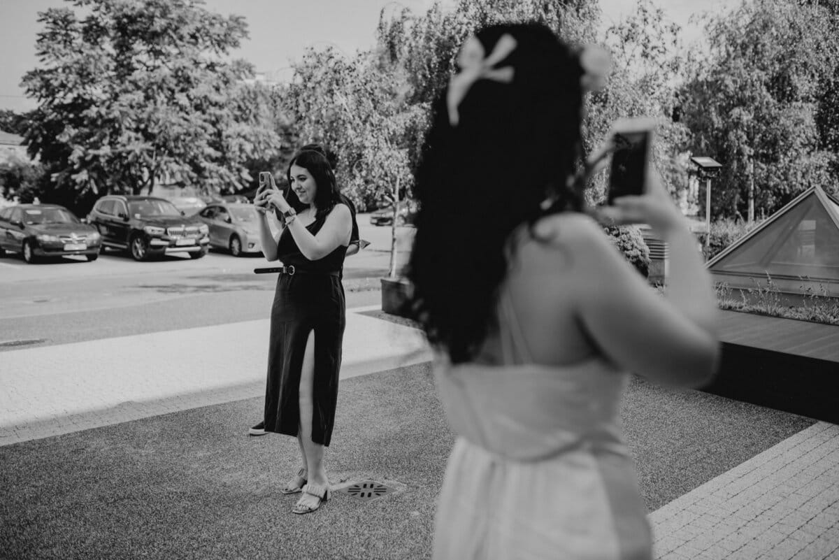 A black and white photo of two women outside, each taking a picture with a smartphone. One woman in a sleeveless dress stands in the background, while the other woman with a flower in her hair, wearing a light-colored dress, stands in the foreground with her back to the camera. The image captures the essence of everyday moments as seen through Marcin Krokowski's eyes.  