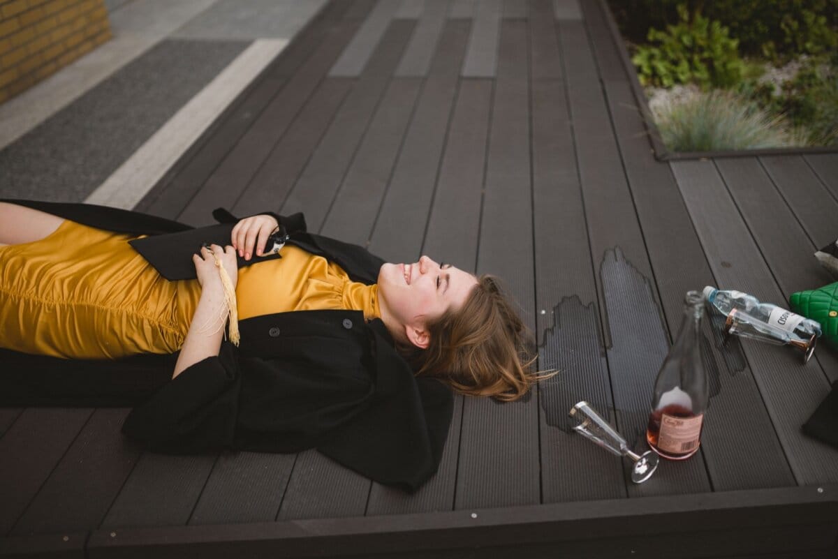 A woman in a yellow dress and black jacket is lying on her back on a wooden deck, smiling and holding a phone. Beside her are two empty bottles, a spilled drink and a glass. Capturing this relaxed and happy moment could easily become one of Marcin Krokowski's event photo shoots in Warsaw.  