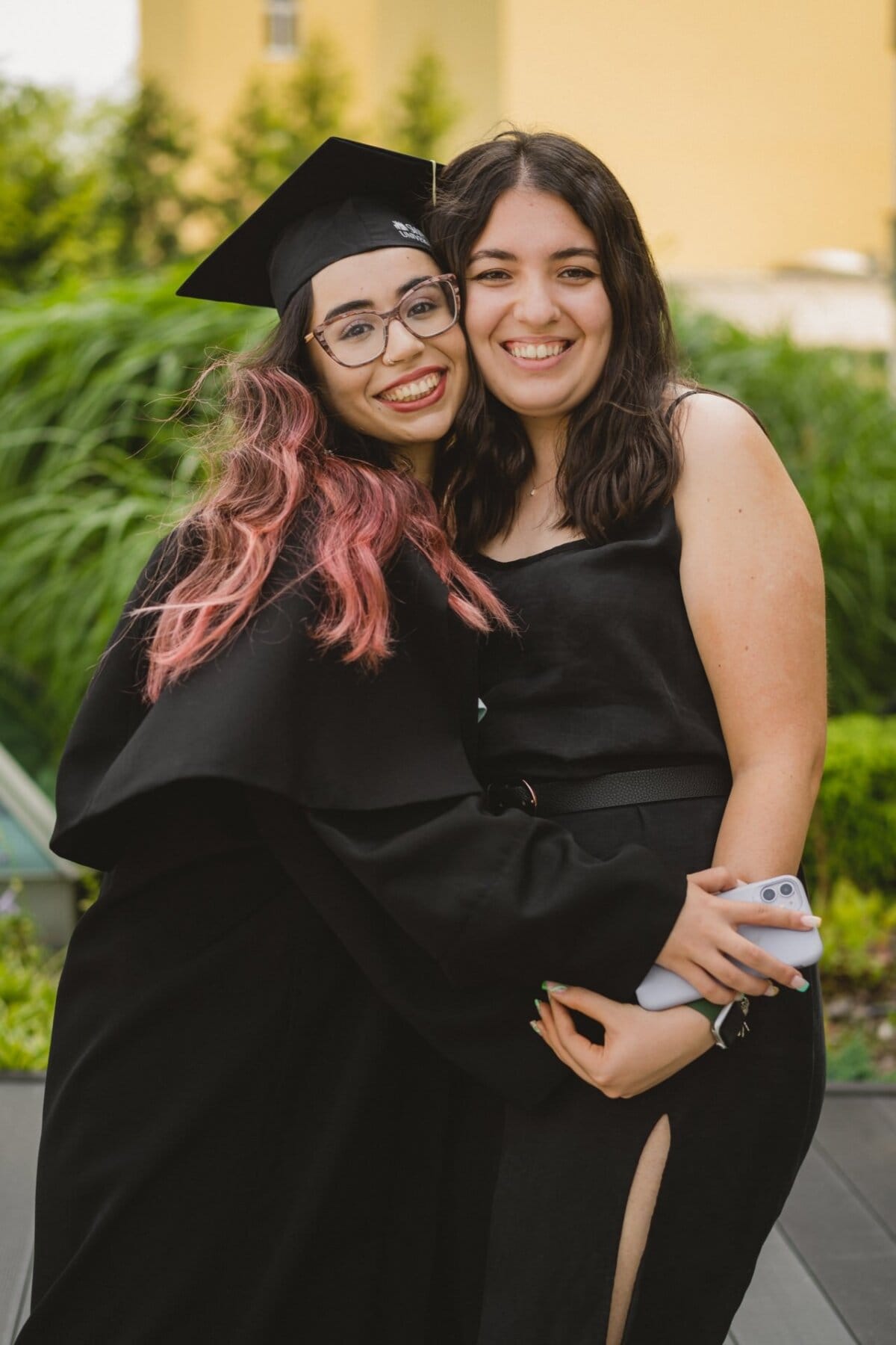 Two women smile and hug each other outdoors during a photo opportunity at the event. One is wearing a black dress and graduation cap and has long dark hair with pink highlights. The other is dressed in a sleeveless black dress and holding a smartphone. Greenery and buildings are visible in the background.   