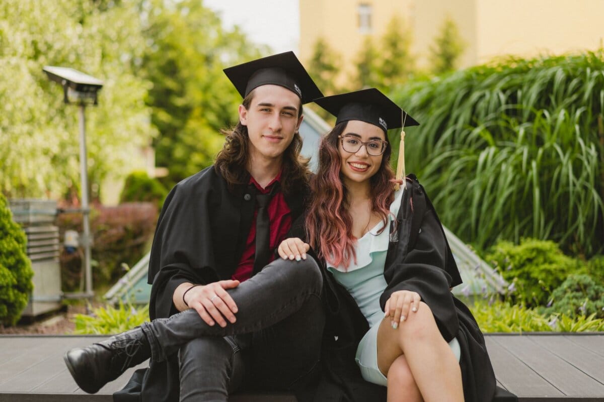 Two people wearing caps and aprons are sitting together and smiling. One has long hair and wears a red shirt and black pants, the other has long hair and glasses and is wearing a blue dress. They are outdoors, with greenery and a building in the background - a beautiful example of event photography.  