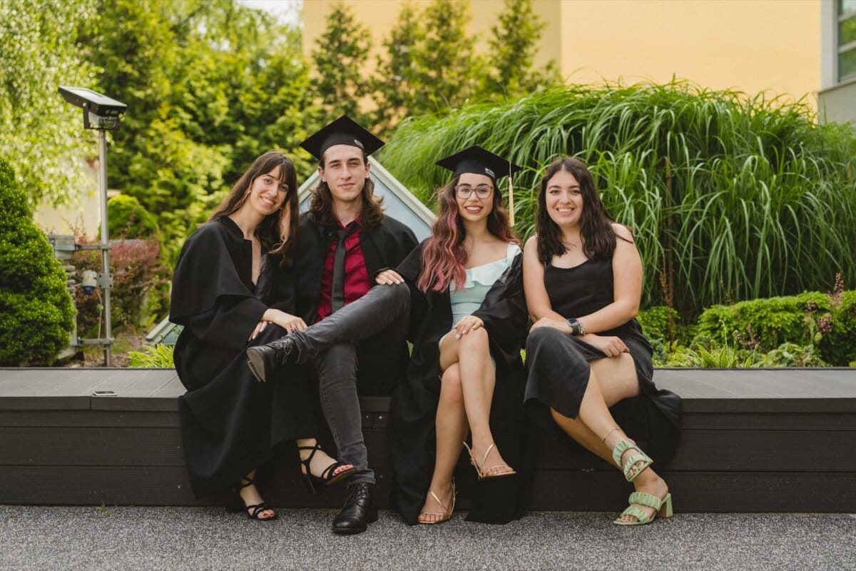 Four young adults sit on a bench and pose for a photo. Three are wearing hats and aprons, and the fourth is wearing casual summer attire. They are outside with the greenery in the background and smiling at the camera. This joyful moment was captured by Marcin Krokowski, event photographer Warsaw.   