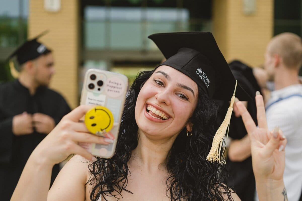 A young woman in a cap and graduation gown smiles broadly as she takes a selfie with her smartphone during a photo recap of events. With her other hand, she holds up a peace sign. In the background, other graduates can be seen mingling.  