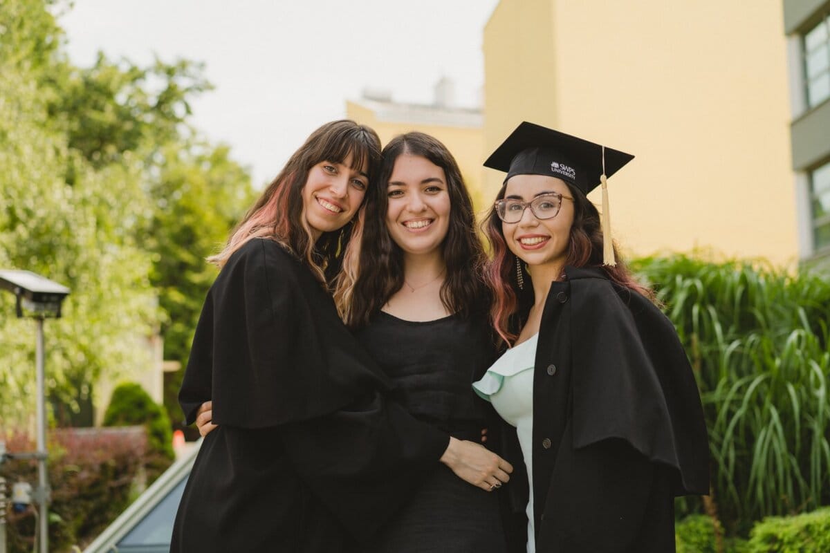 Three young women are smiling and standing close together outside on a sunny day. They are dressed in black graduation gowns, one of them wearing a mortarboard cap. They appear to be celebrating their graduation. The greenery and building in the background, captured by Marcin Krokowski, event photographer warszawa.   