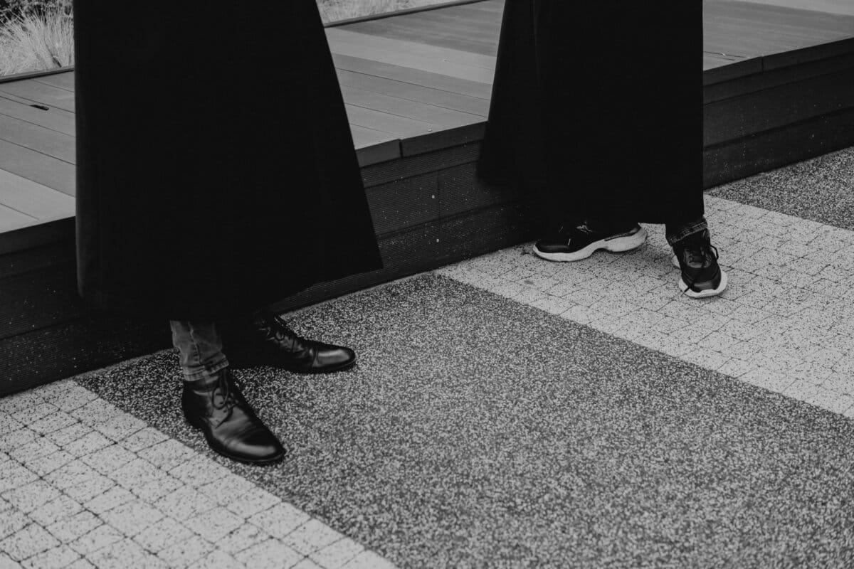 Black and white photo showing two people standing next to a wooden platform. Only the lower parts of the legs and shoes are visible - one is wearing smart shoes with jeans, the other sports sneakers with long pants. Both are wearing long, dark clothes that partially cover their legs, which epitomizes the style of event photography.  