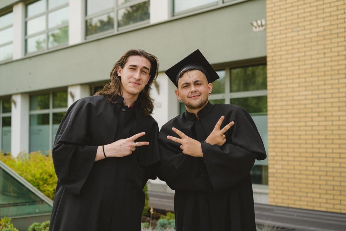 Two graduates dressed in black gowns stand in front of the building, smiling and making peace signs with their hands. One of them is wearing a graduation cap. In the background are windows and a brick wall. This moment was beautifully captured by Marcin Krokowski, a talented event photographer Warsaw, during the photo coverage of the events.   