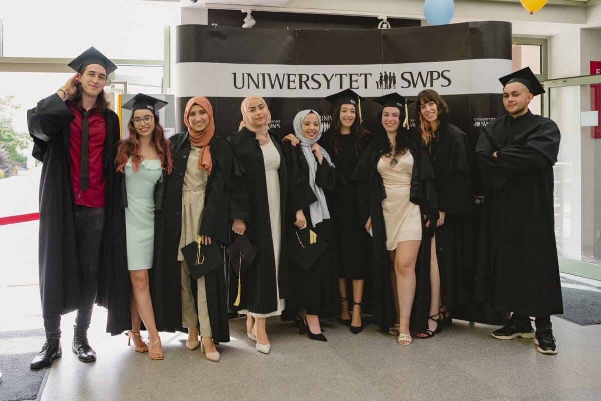 A group of nine graduates wearing togas and birettas pose for a photo against the backdrop of the "SWPS UNIVERSITY" sign in black. Four women are wearing hijabs. Some are holding diplomas, and all are smiling and look happy, captured by event photographer Marcin Krokowski.  