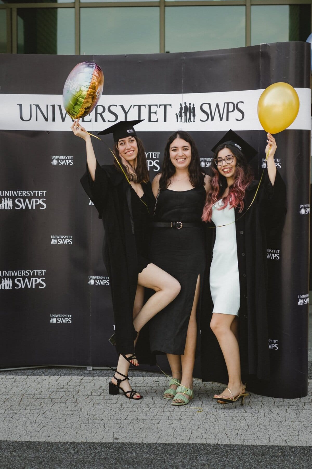 Three women in graduation gowns and caps pose with balloons in the background of "SWPS UNIVERSITY." The women are smiling and celebrating their accomplishment, with one holding a multicolored balloon and the other holding a yellow balloon - a perfect photo opportunity to capture a joyful moment at the event. 