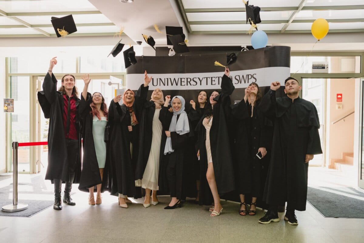 A group of students in graduation attire celebrate by tossing their caps up inside the building with a banner reading "SWPS University." They stand in front of the entrance, smiling and cheering, the joy of their achievement captured in a photo report of the events. 