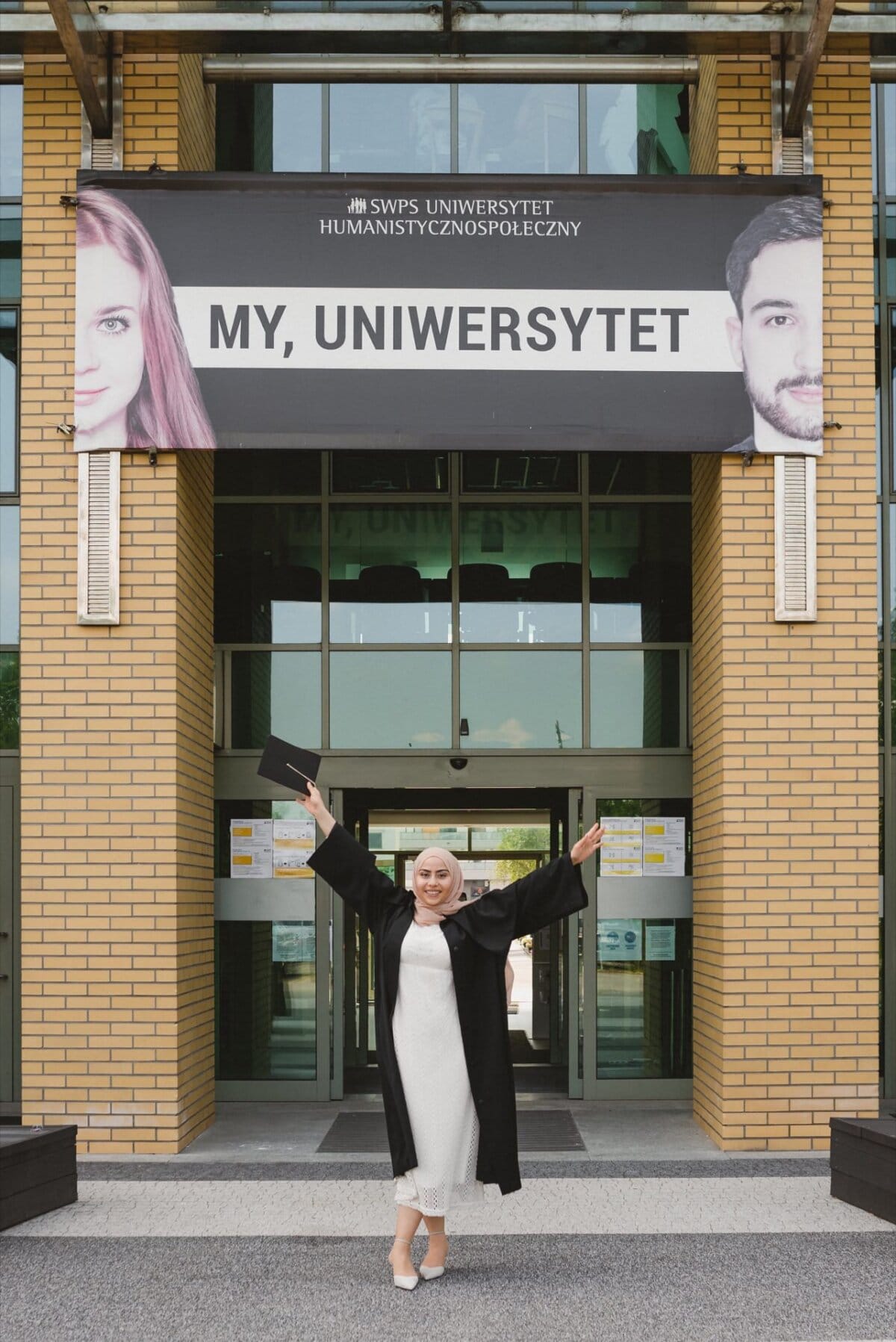 A smiling graduate in a hijab and black dress holds her diploma in front of the SWPS University of Humanities and Social Sciences building. A banner at the entrance reads "MY, UNIVERSITY" and the faces of a man and a woman, beautifully captured by event photographer Marcin Krokowski. 