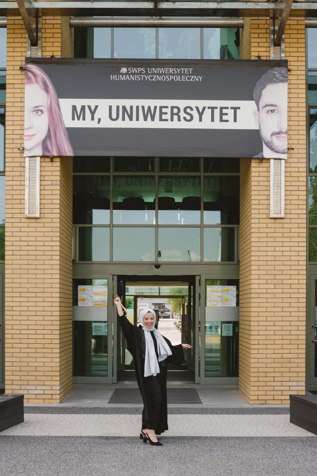 A person dressed in high school graduation attire stands joyfully with arms outstretched in front of the university building. Above the entrance, a banner with images of two faces with the words "MY, UNIVERSITY" and "SWPS UNIVERSITY OF HUMANISM". This moment was beautifully captured by event photographer Marcin Krokowski.  