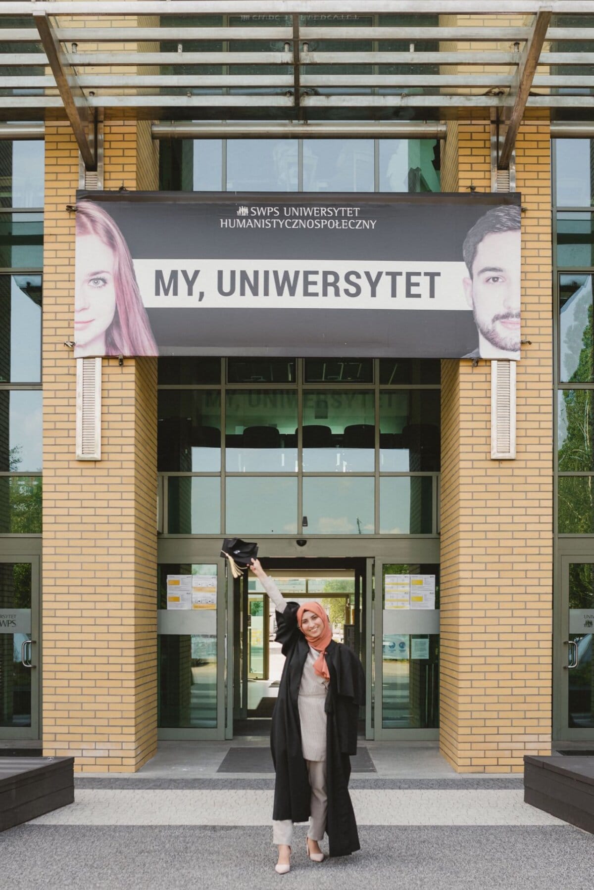 A woman in a graduation gown and hijab stands joyfully holding a graduation cap in front of the entrance to SWPS University. Above the entrance, a banner with the words "MY, UNIVERSITY" and images of a woman and a man on either side, captured perfectly for a photo essay of the event. 