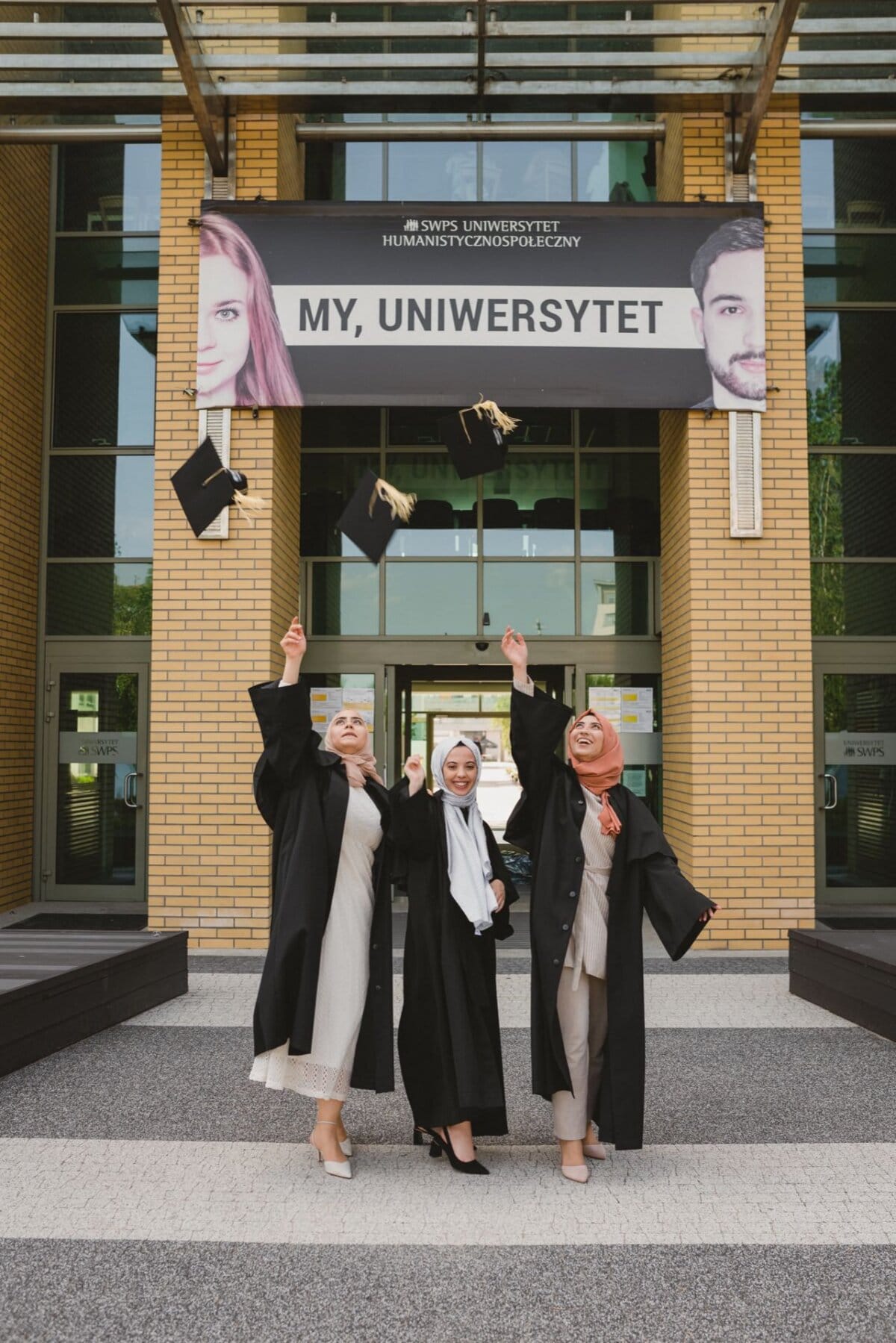 Three women wearing graduation togas throw their birettas in the air in front of the university building, which bears the inscription "MY, UNIVERSITY." They stand on the paved area, smiling and celebrating their achievement, perfectly captured by an event photographer Warsaw specializing in event photography. 