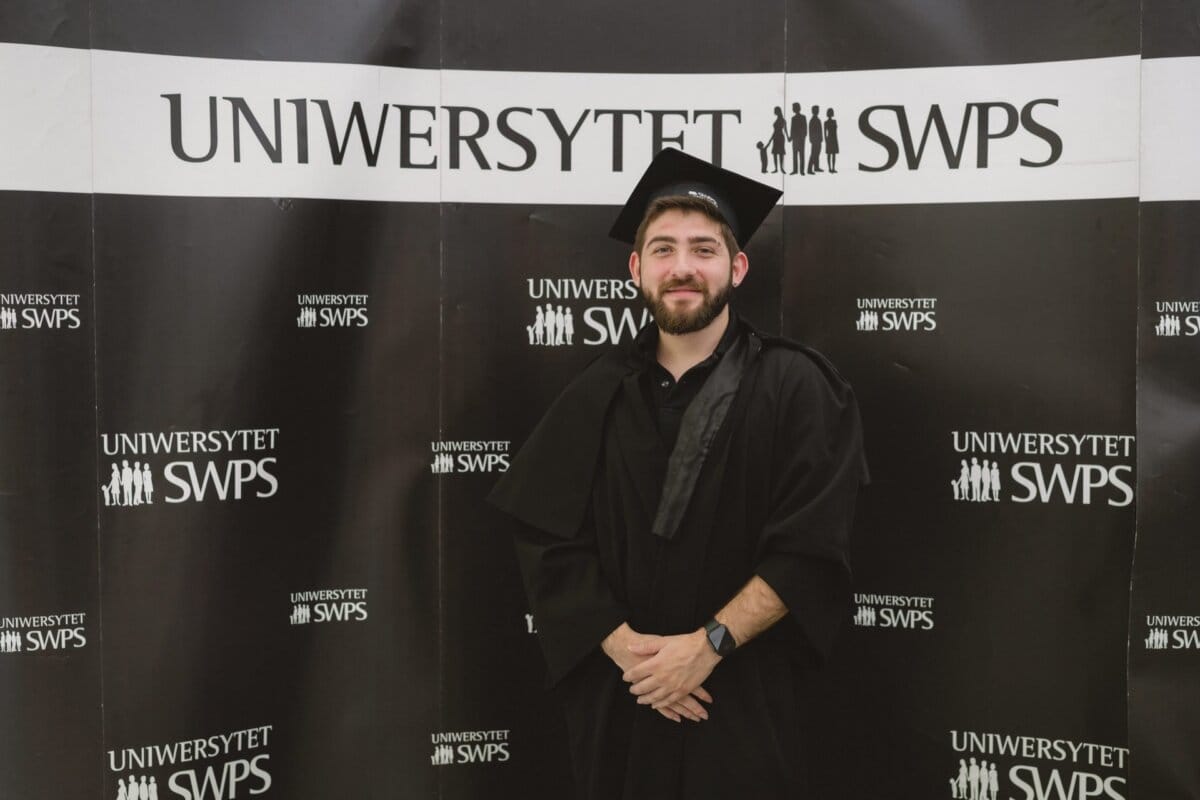 A person wearing a graduation cap and toga stands against a background with repeated "SWPS UNIVERSITY" logos. The figure is smiling and posing with his hands folded in front, which was beautifully captured by the event photography. 