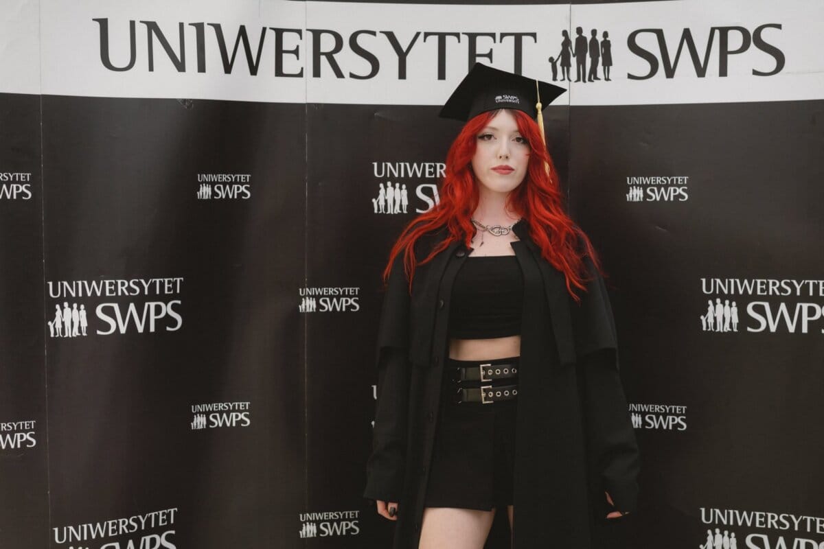 In the background "SWPS UNIVERSITY" stands a young person with long red hair. They are wearing a black graduation cap, a black sleeveless top and a black skirt belted with a wide belt. They have a serious expression on their faces and pose confidently, which was captured perfectly by event photographer Warsaw.  