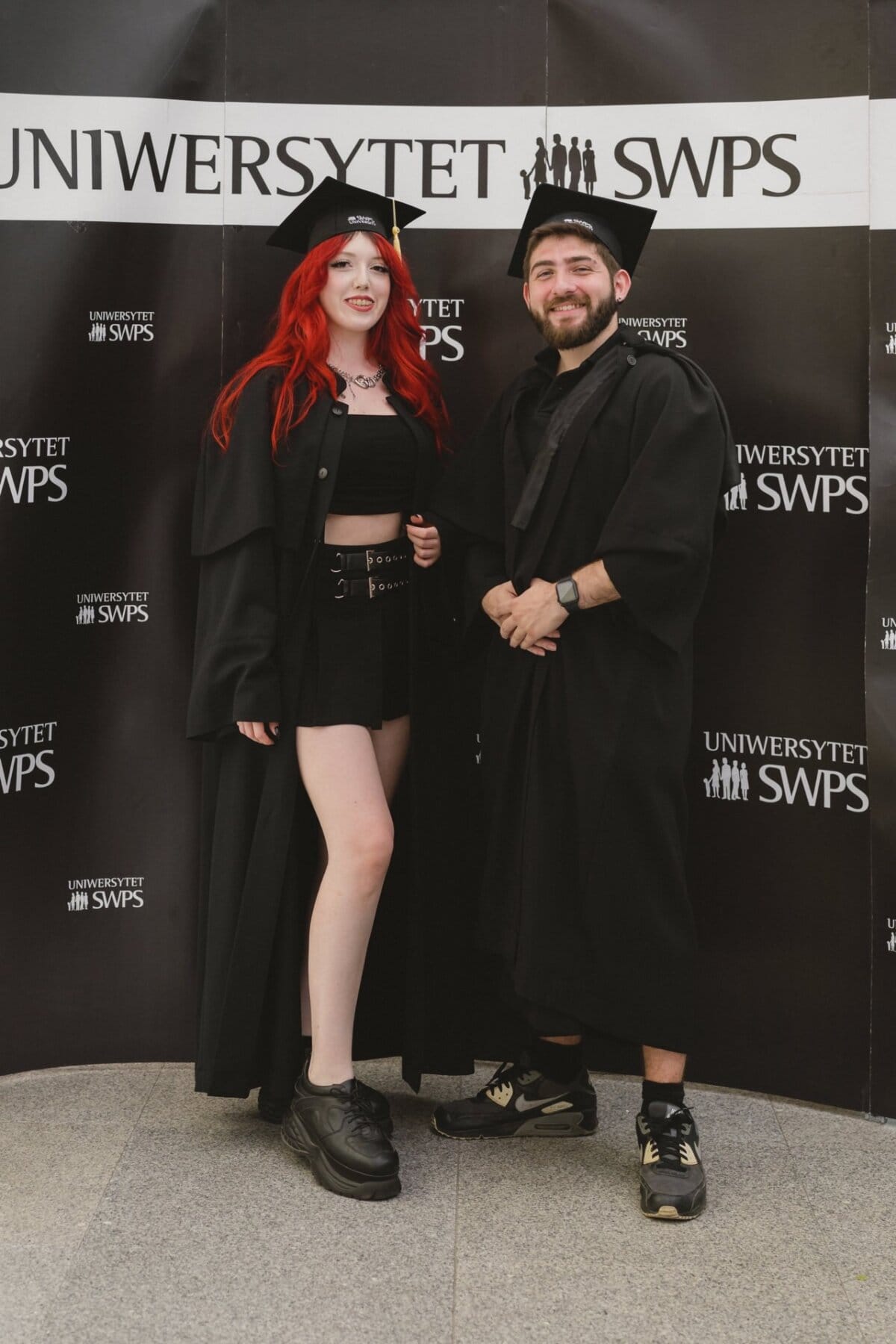 Two people dressed in black togas and graduation caps stand smiling in front of a background with the repeated inscription "SWPS UNIVERSITY." One has red hair, a dress and platform shoes, while the other has a beard and wears a black t-shirt and sneakers. This event photo captures the joy of this special day.  