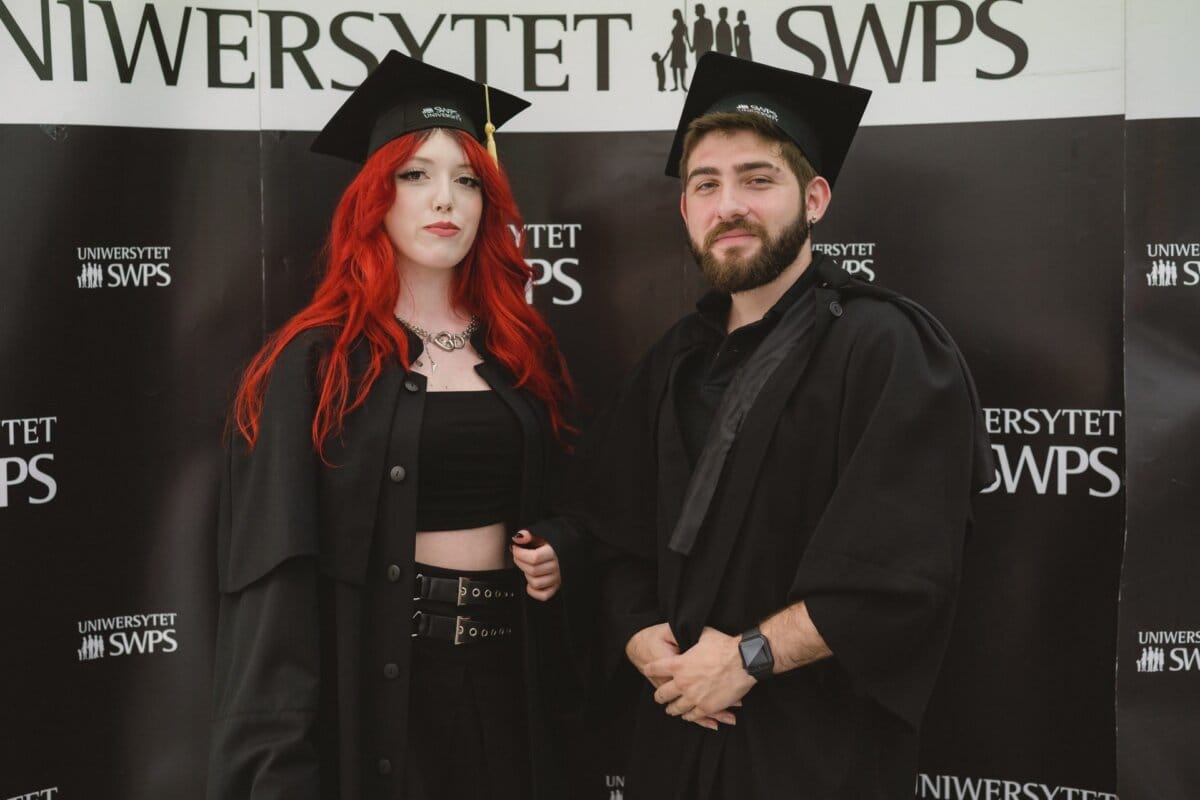 Two graduates pose with the SWPS University logo in the background. Both are dressed in black graduation gowns and caps. The graduate on the left has long red hair and wears a choker, while the graduate on the right has a beard and mustache, which is beautifully captured in this event photo.  