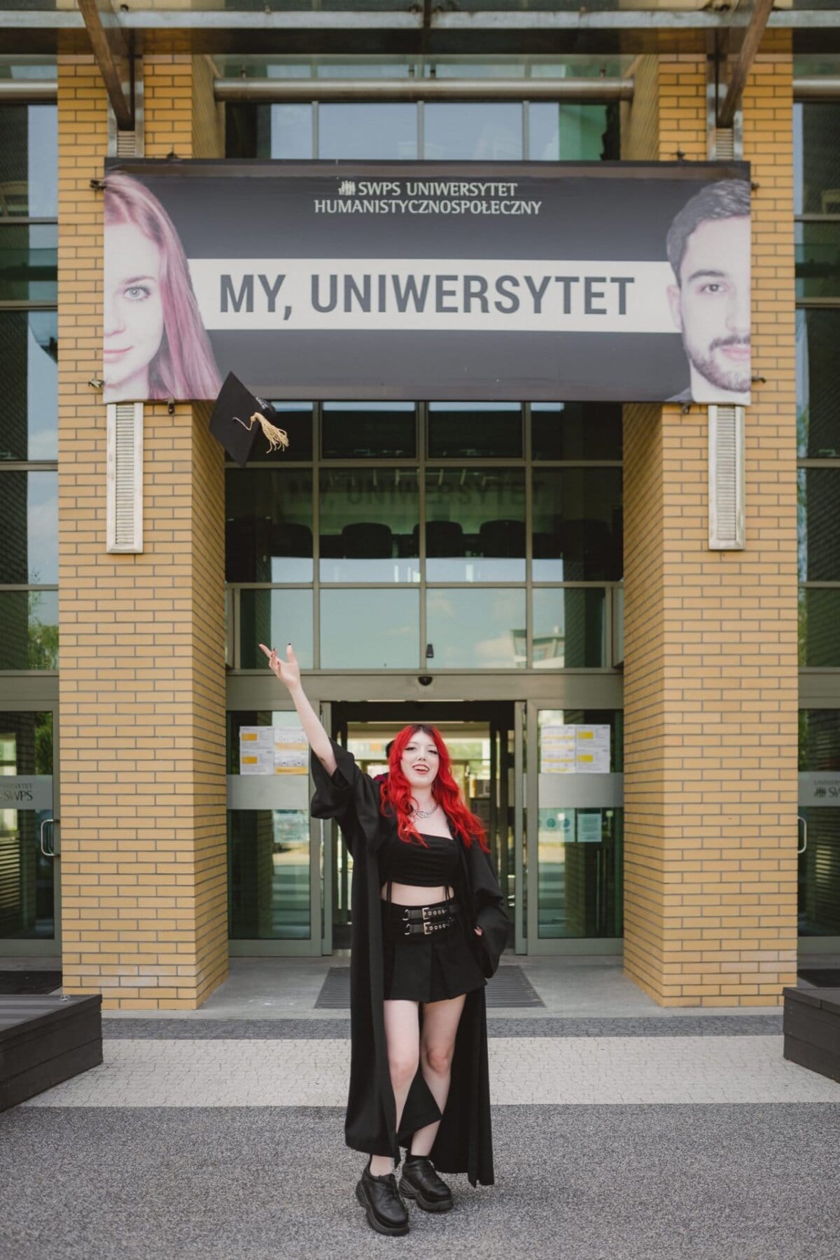 A student with long red hair, dressed in a graduation gown and black outfit, joyfully tosses her graduation cap up in the air, standing in front of a building with a large banner reading "MY, UNIVERSITY" and depicting two faces - beautifully captured by the image of the event.