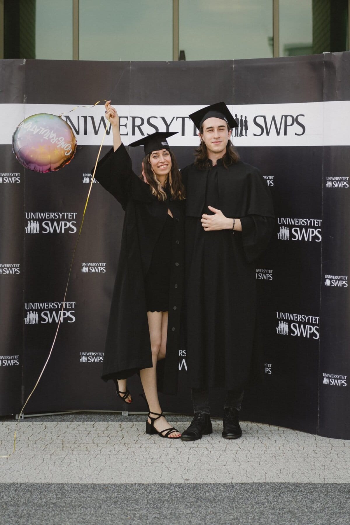 Against the background of "SWPS UNIVERSITY" stand two people, both wearing black graduation gowns. One holds a colorful balloon and smiles widely. The other puts his hand on his chest and looks into the camera. This photo captures a joyful moment from the event's photo essay.   