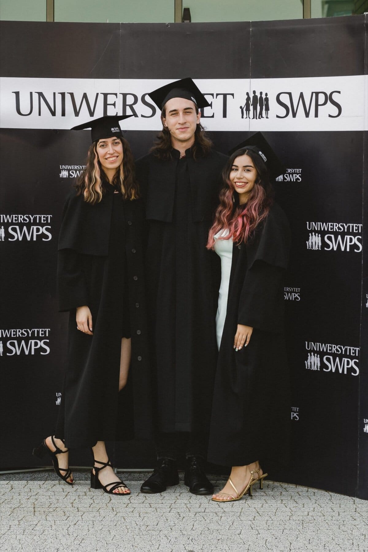 Three graduates in black aprons and mortarboards stand together, smiling, in front of a backdrop that reads "SWPS UNIVERSITY." The middle person is taller and stands between the other two people, who are wearing high heels. This event photography perfectly captures the joy of their achievement.  