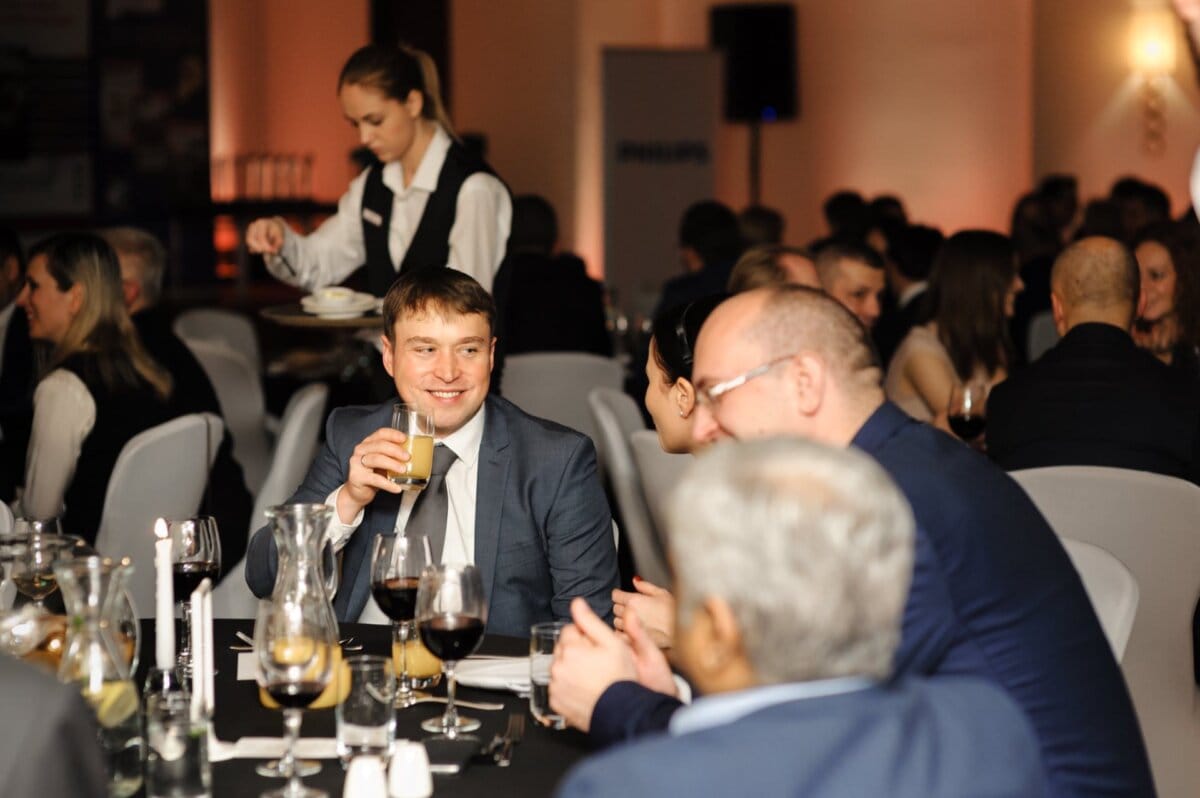 Man at table smiles while holding juice and looks at other guests