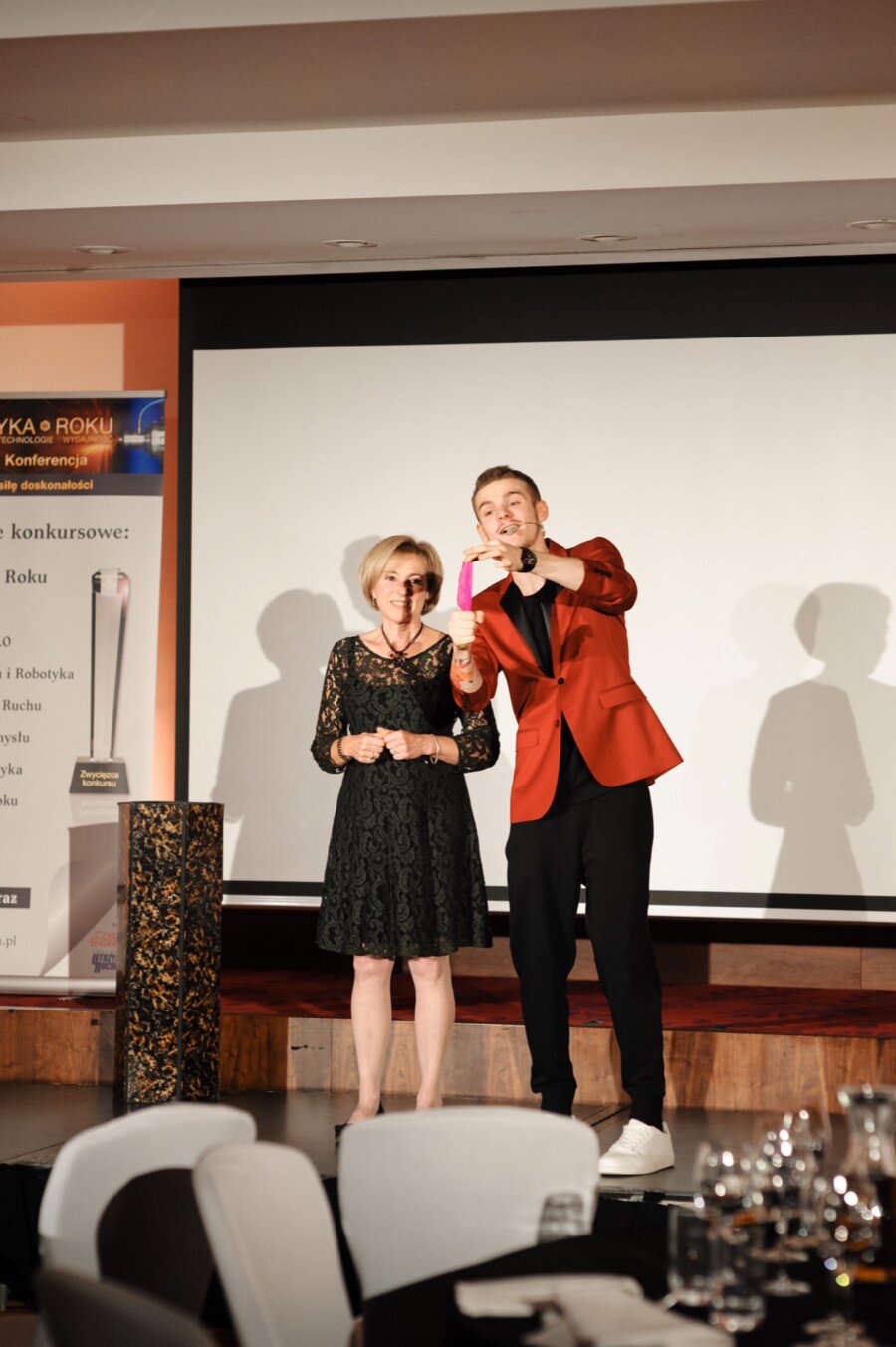 A woman in a black dress and a man in a red jacket stand on stage