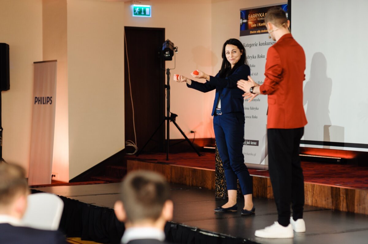 Woman on stage with red balls in her hands looks has man in red jacket