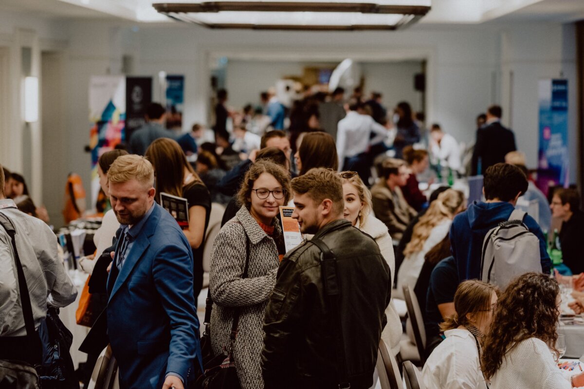 A large group of people is gathering in a spacious, well-lit conference room. They are engaged in conversation, some standing, others sitting around tables. The atmosphere seems busy and sociable, perfect for event photography Warsaw, where attendees are networking and interacting with each other.  
