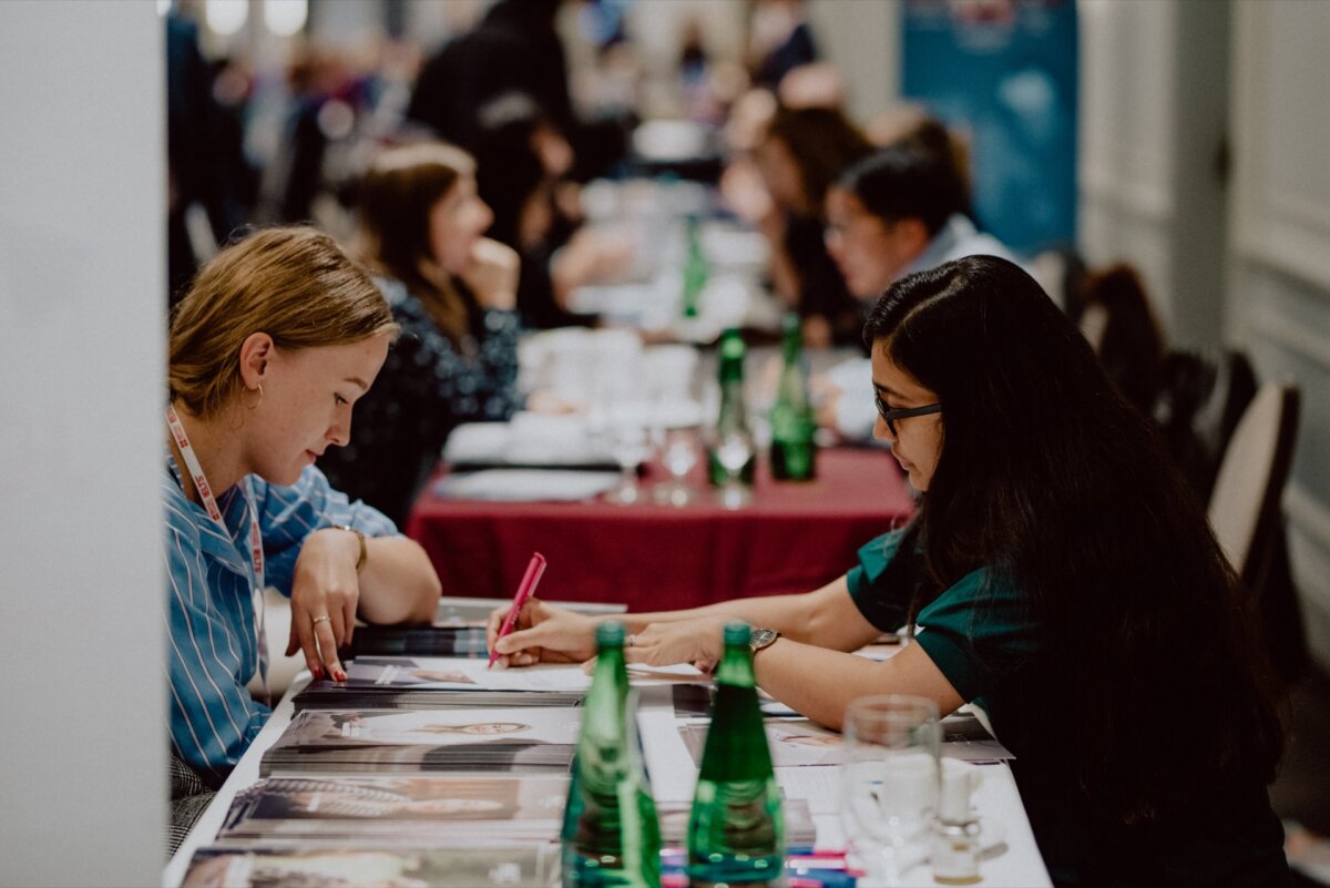 Two women sitting at a table are discussing, one is writing on a piece of paper. Various people are sitting at other tables in the background, water bottles and glasses are visible. The place appears to be a conference or networking event, ideal for photo coverage of events.  