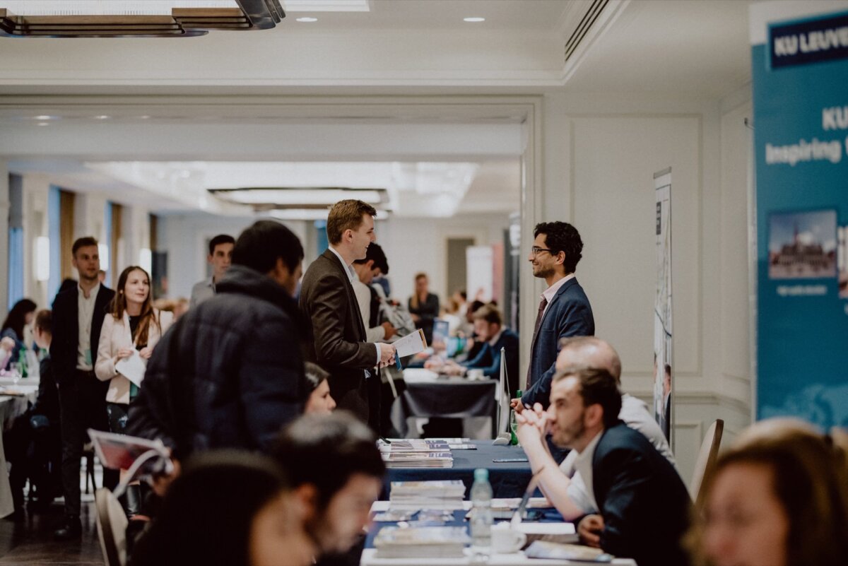 People attending a busy career or education fair in a spacious, well-lit room. Various booths with informational materials and representatives contacting visitors are visible. Two men in business attire are talking in the foreground, which is perfect for capturing a photo essay of the events.  