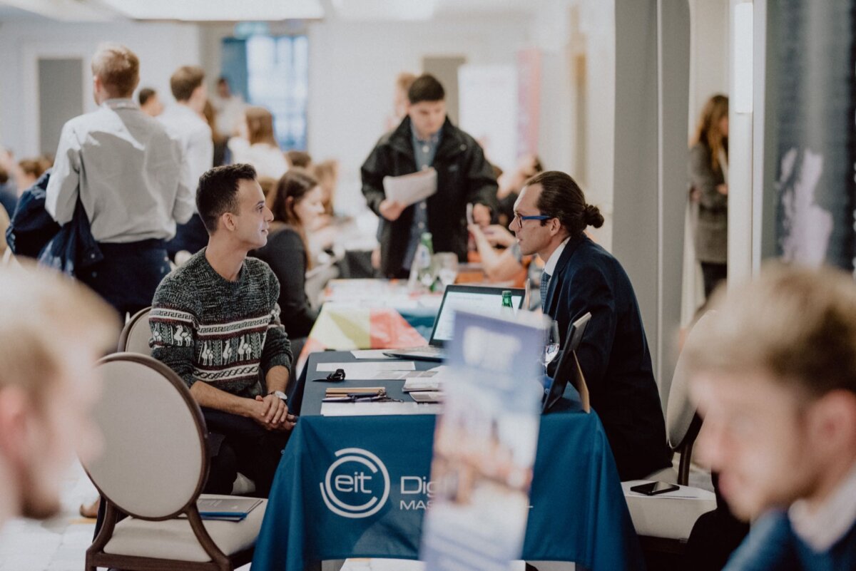 A busy job fair in a well-lit area. Participants, both seated and standing, are engaged in discussions at various booths. One person in a sweater talks to another person seated behind a table decorated with materials and a banner reading "EIT Digital," creating the perfect setting for event photography.  