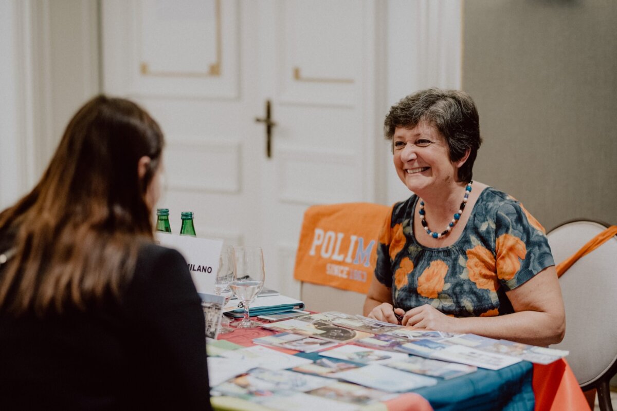 A woman with short hair, wearing a floral dress and a beaded necklace, sits at a table strewn with photos and flyers representing event photography. She smiles and talks with another woman with long hair across from her. A banner with the words "Polim..." hangs above the chair.  