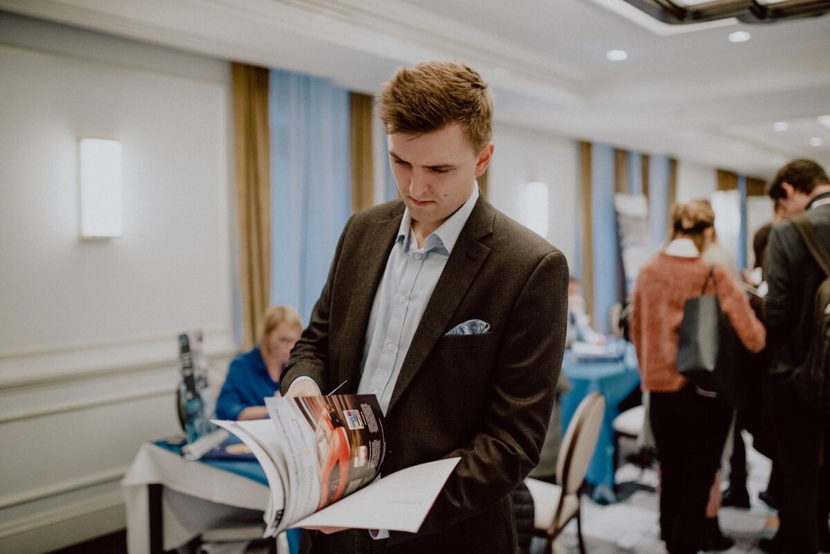 A young man in a dark jacket and light blue shirt stands in a well-lit room, holding and reading a brochure. In the background, other people can be seen interacting, capturing the essence of a "photo event," indicating a professional or networking event. 