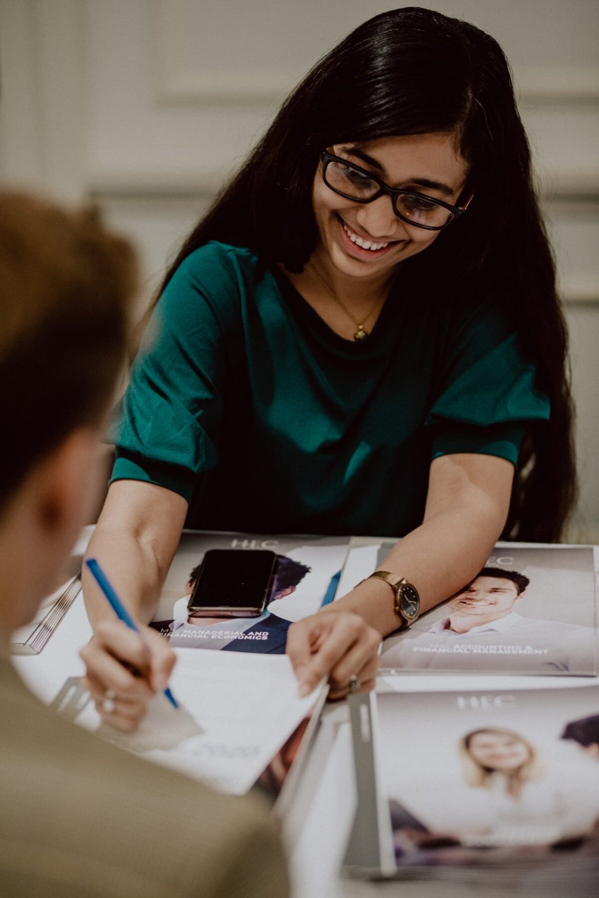 A person with long dark hair and glasses smiles, writing on a piece of paper. They are sitting at a table, with brochures and a cell phone in front of them. The person, dressed in a green shirt, gives the impression of being busy in conversation, perfectly capturing the essence of event photography.  