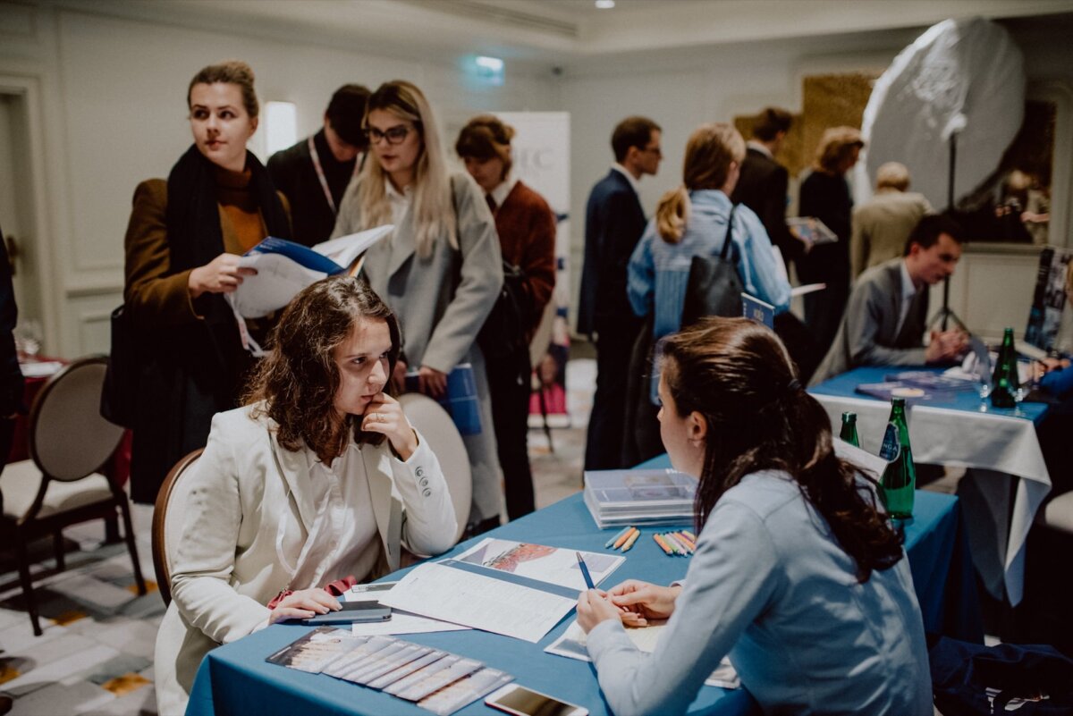 A crowded room where people are having conversations and holding documents at various tables. In the foreground two women are sitting at a blue table, one seems to be listening attentively, the other is talking. Various brochures and newspapers are spread out on the table - a perfect scene for event photography by a talented event photographer Warsaw.  