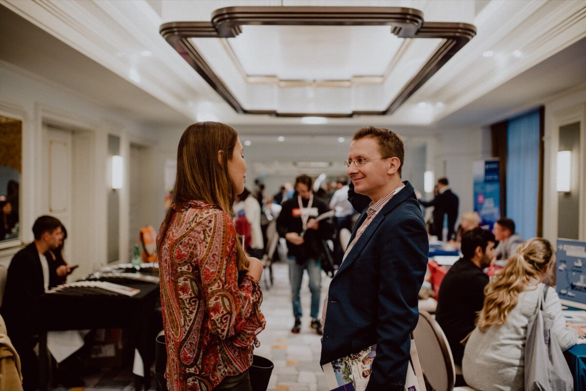 Two people are facing each other, busy in conversation, in a bustling room with people in the background. The man is dressed in a suit and the woman in a patterned blouse. The setting suggests a professional event or conference, beautifully captured in an event photography session.  