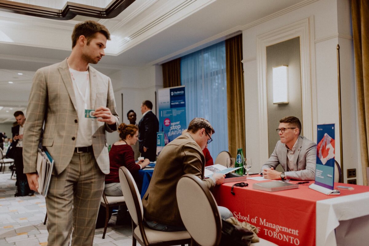 A man in a beige suit holds a drink and walks past tables where people are sitting busy discussing. On one table is a banner of the University of Toronto's School of Management. The setting is reminiscent of a professional event or career fair, as beautifully captured by event photography.  